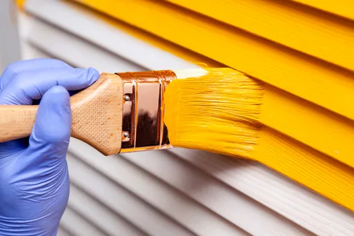 Gloved hand brushing bright yellow paint onto lap siding